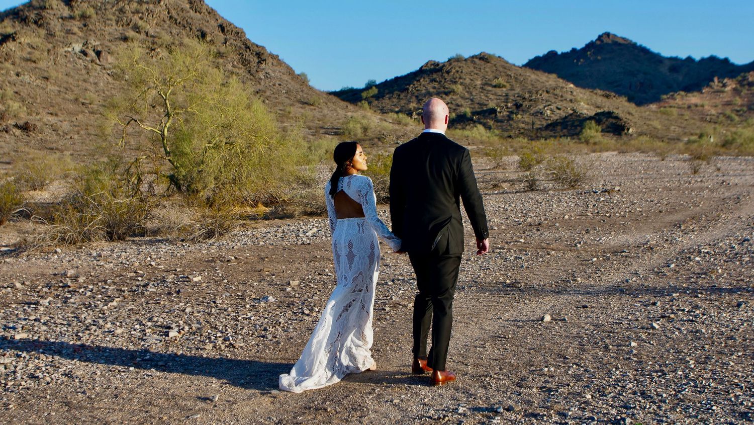 Two people in formal attire walk together on a desert path with rugged mountains in the background.