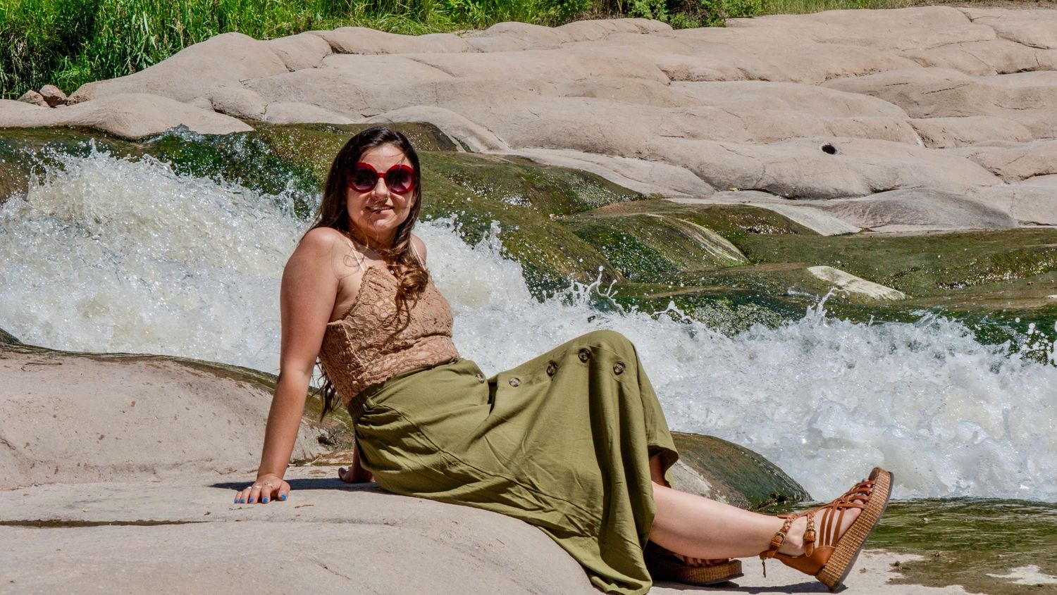 Someone in a green skirt and brown top sits casually by rushing water on smooth rocks on a sunny day.