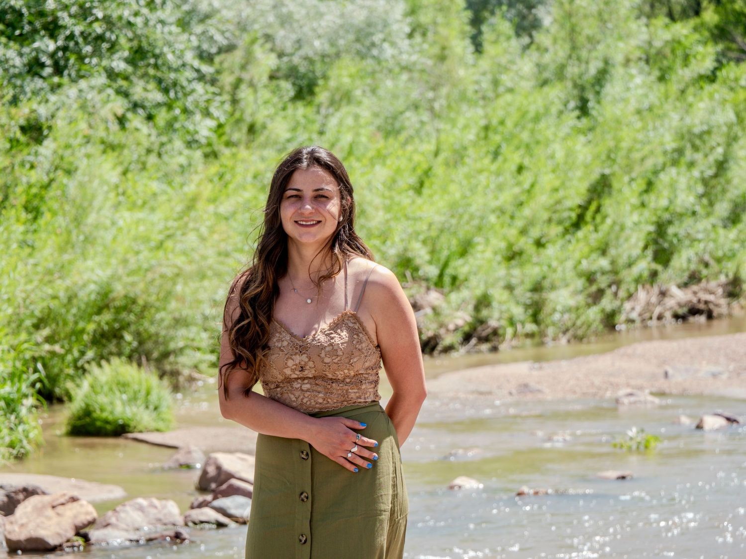 Woman in olive green dress poses by sparkling river on sunny summer day.