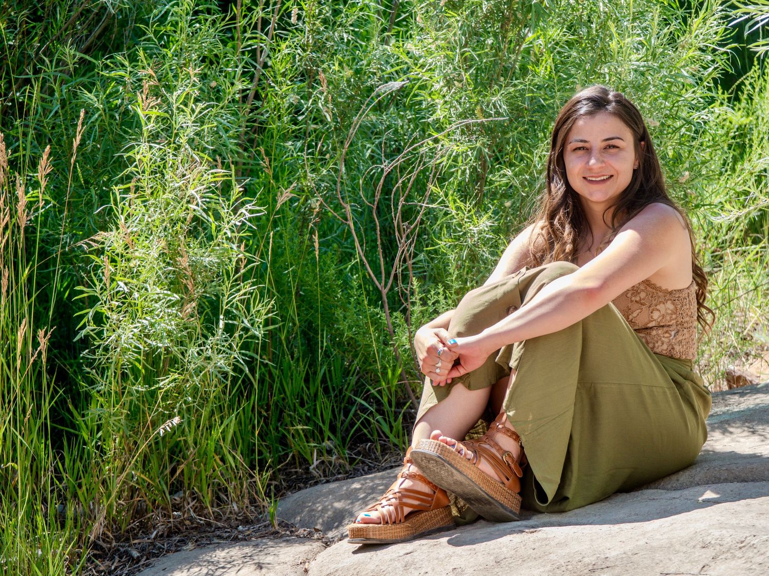 Woman in olive green dress and cowboy boots poses outdoors in natural setting with tall grass.