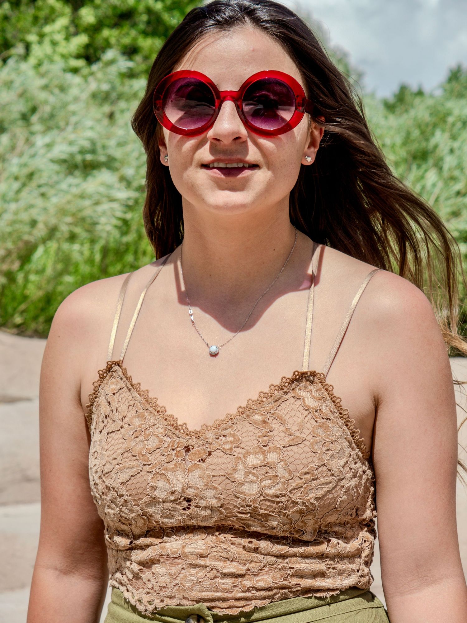 A person in a floral dress and red sunglasses enjoys a sunny day outdoors with a joyful expression.