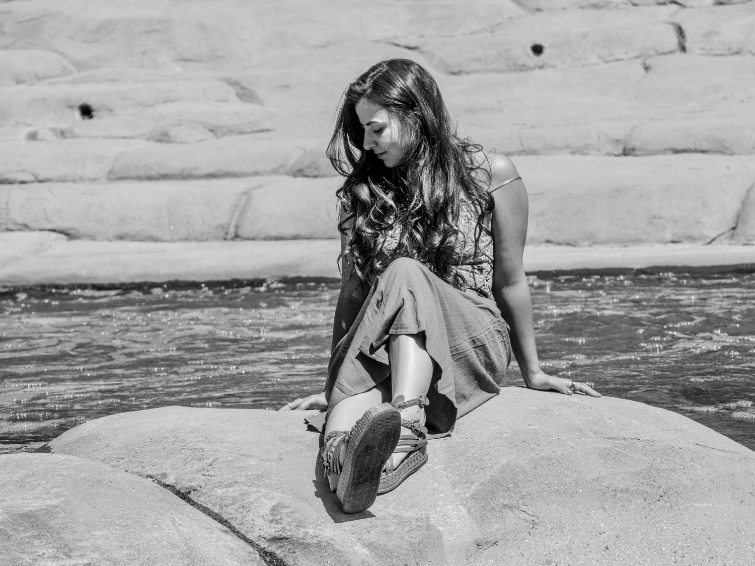 Vintage black and white photograph showing someone sitting on rocky shoreline near water in casual dress.