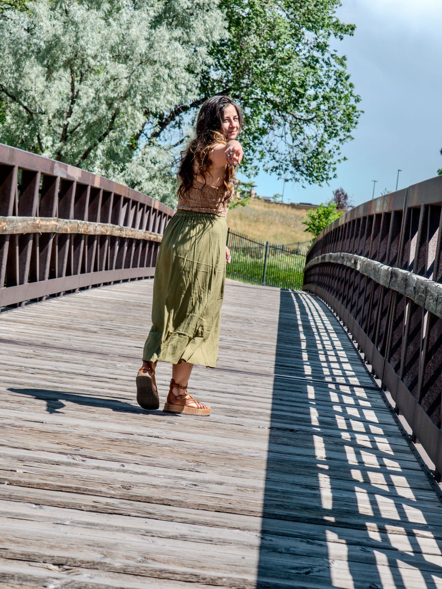 A person in a green dress walks along a wooden bridge boardwalk with metal railings on a sunny summer day.