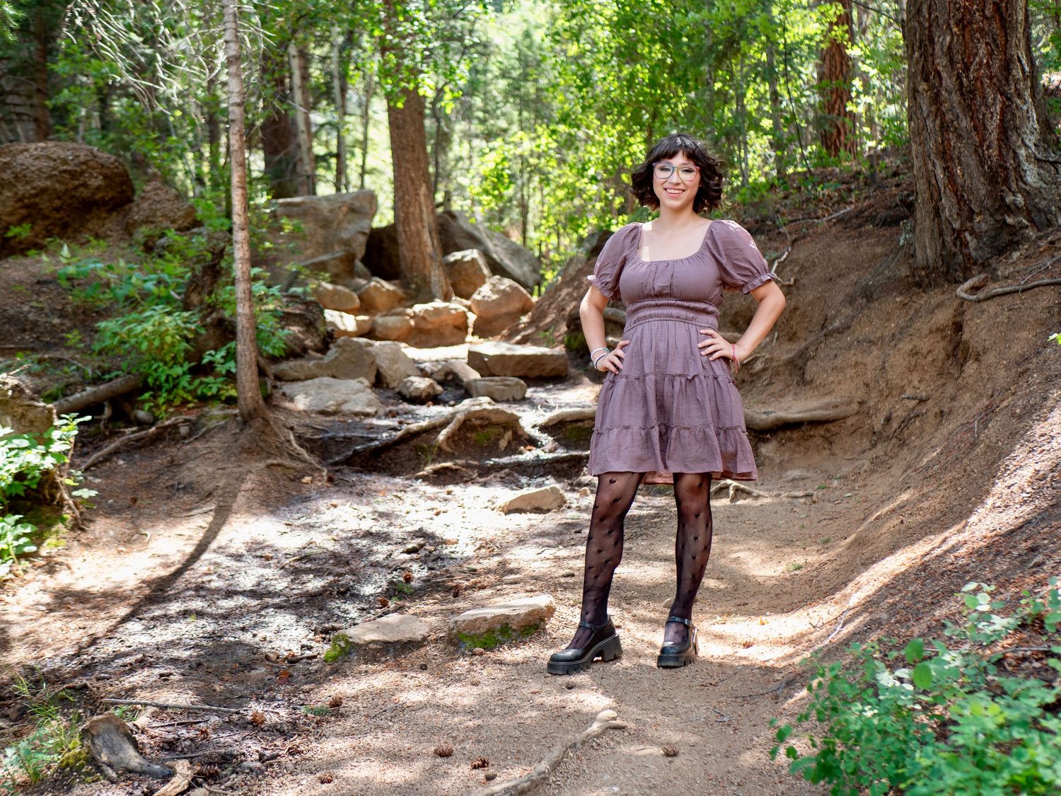 A hiker stands on a scenic forest trail lined with tall trees and rocks in dappled sunlight.