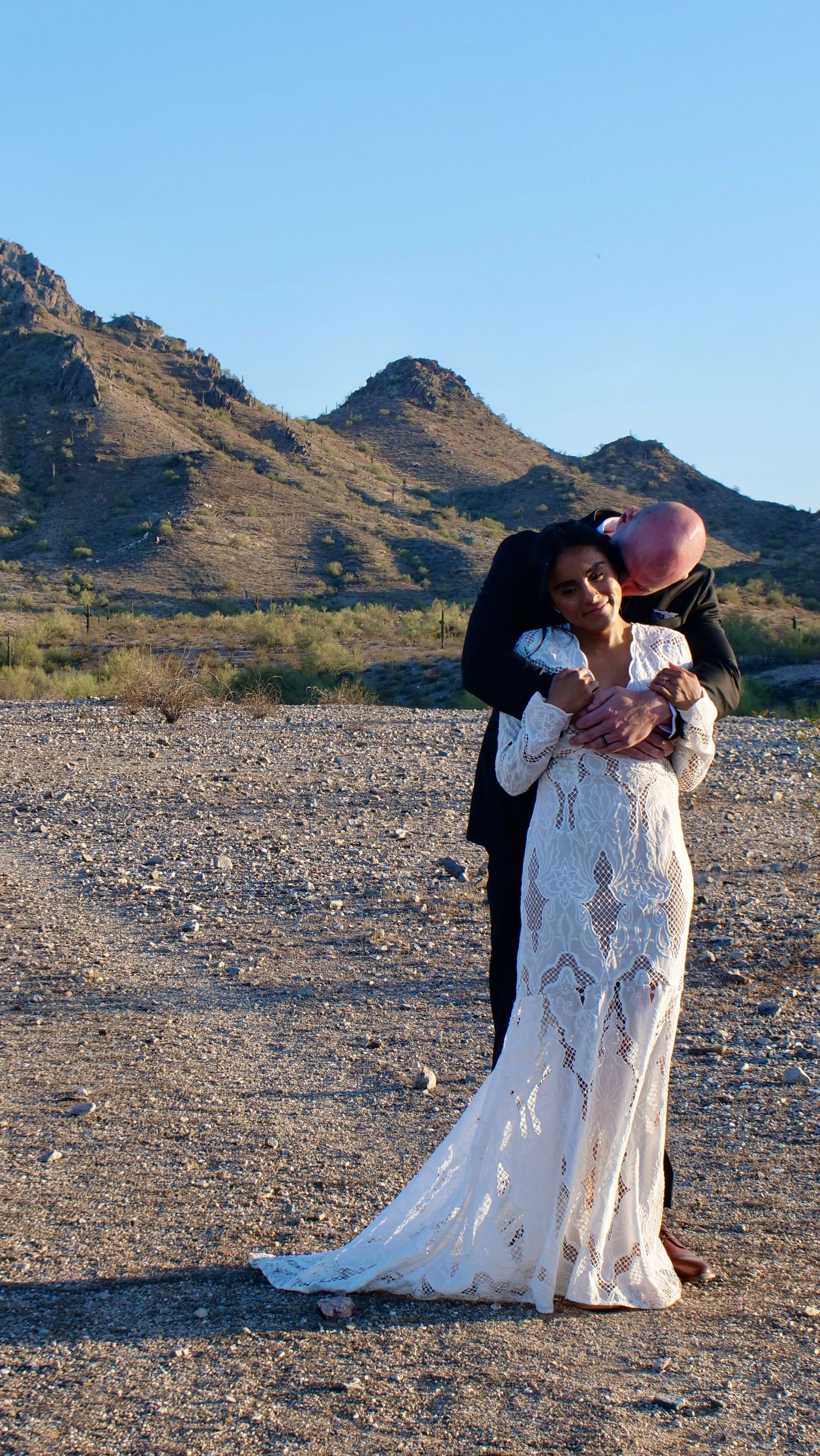 A romantic desert wedding photoshoot shows a couple embracing in front of rocky mountains during sunset.