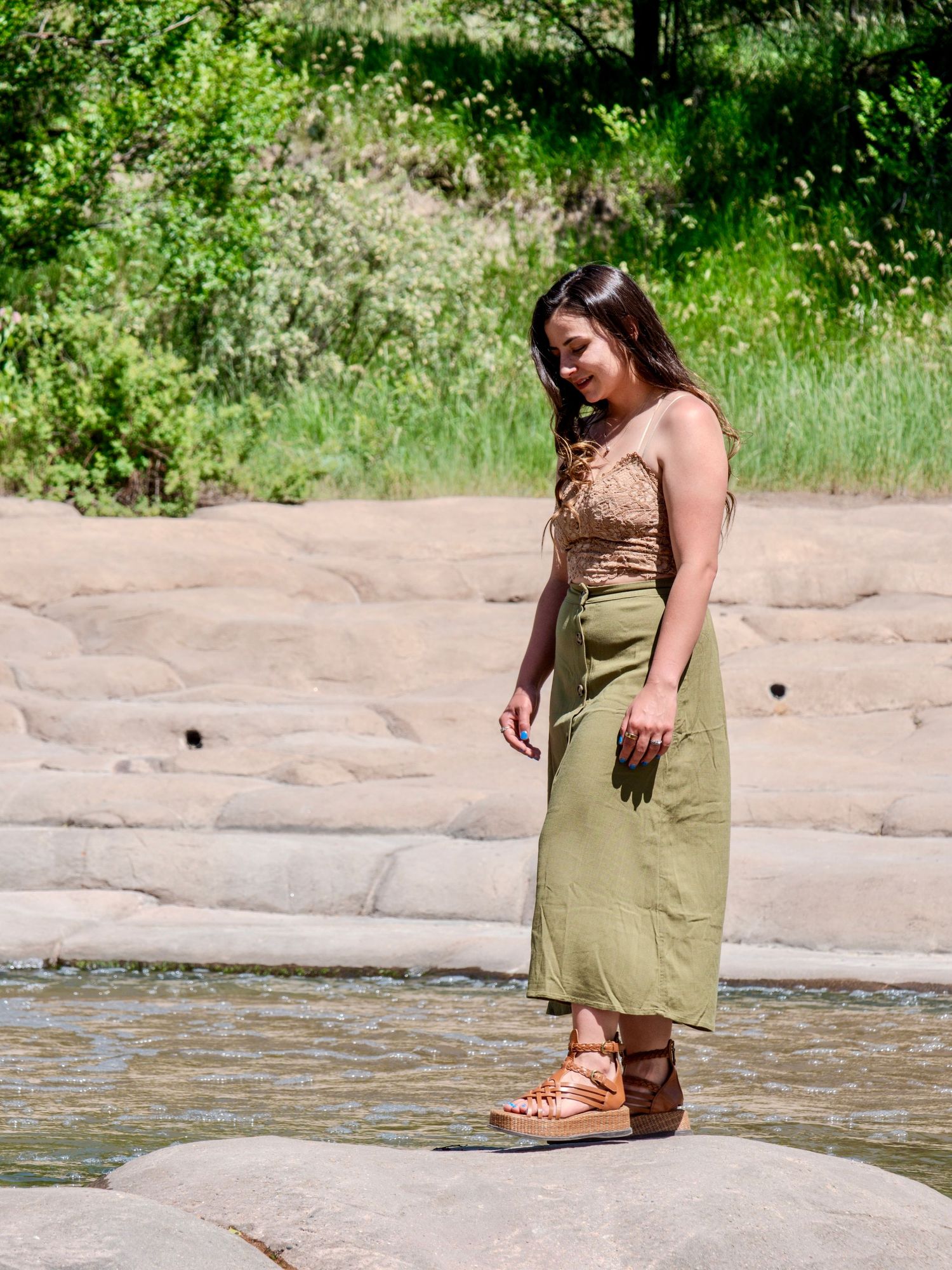 Someone wearing a sage green dress stands near flowing water on rocky terrain on a sunny summer day.