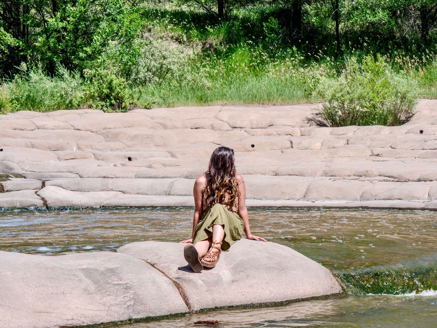 Person sunbathing on a large flat rock by a flowing river on a sunny summer day.