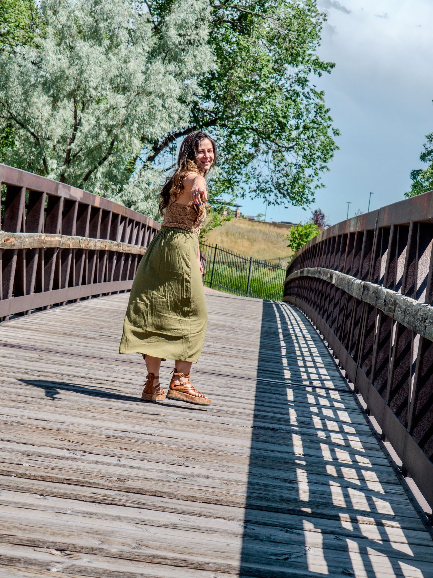 A person in an olive dress walks across a wooden bridge pathway surrounded by trees and greenery on a cloudy day.