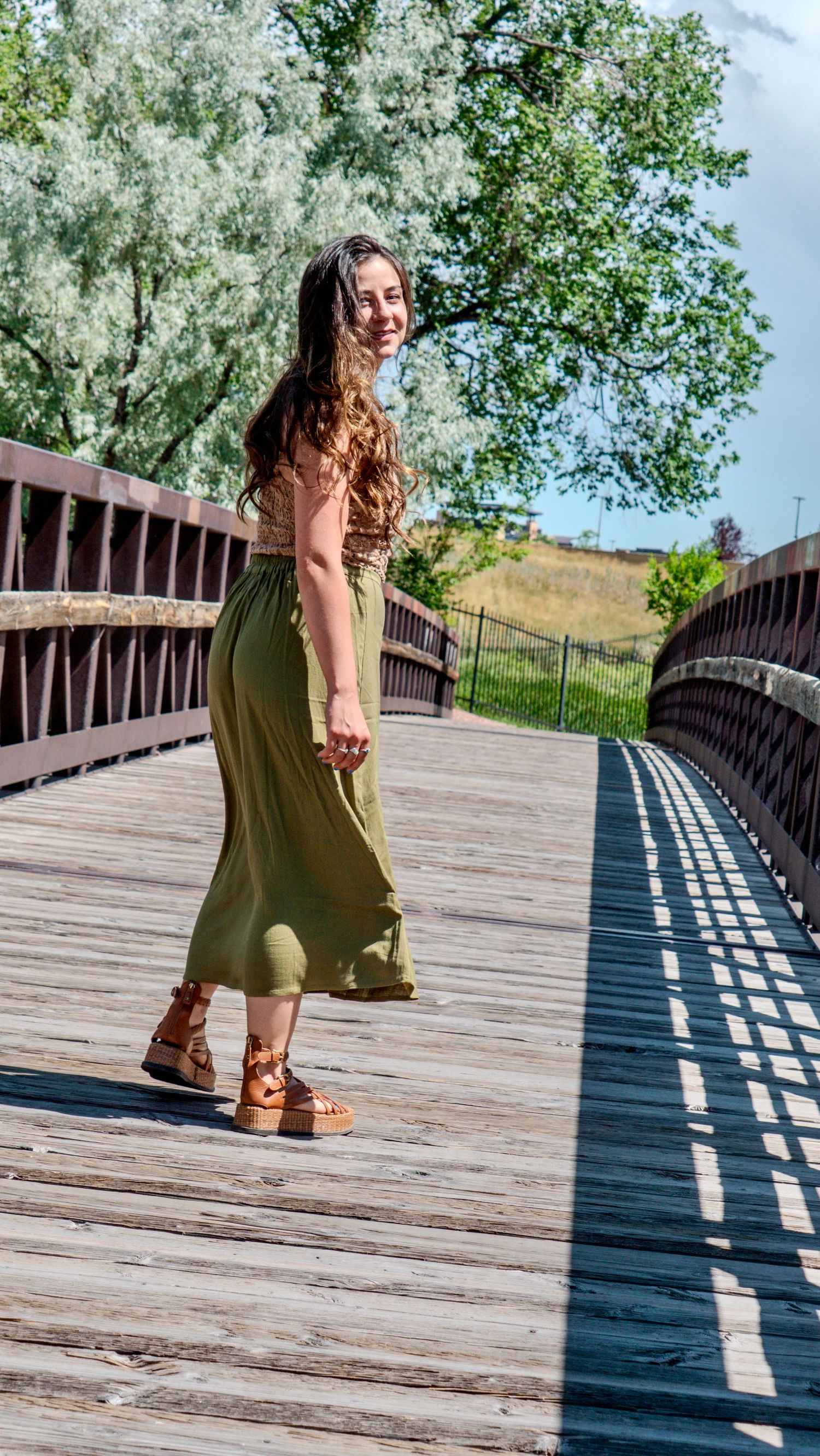 Person in flowing olive skirt twirls on wooden bridge with lush green trees in background.