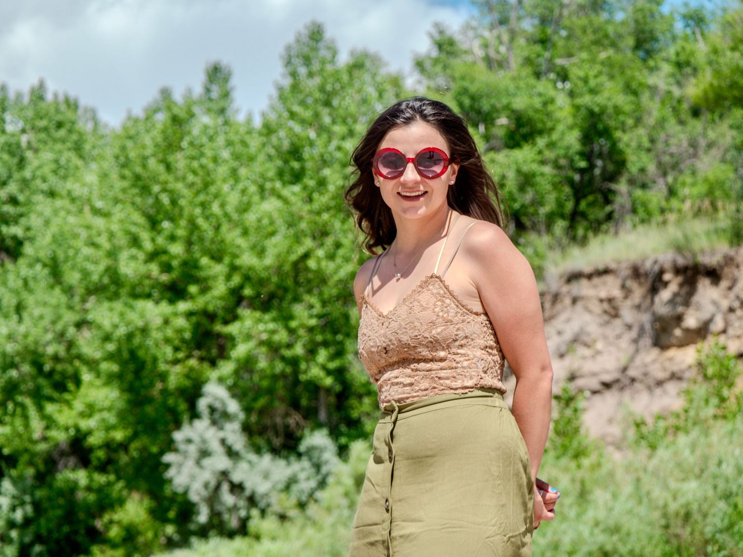 A person in a beige halter top and high-waisted skirt poses outdoors on a sunny day wearing red sunglasses.