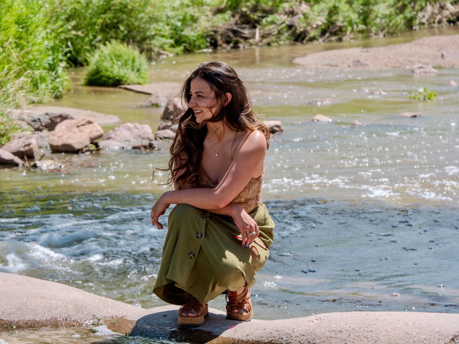 Person in casual attire sitting thoughtfully by flowing stream on rocky terrain.