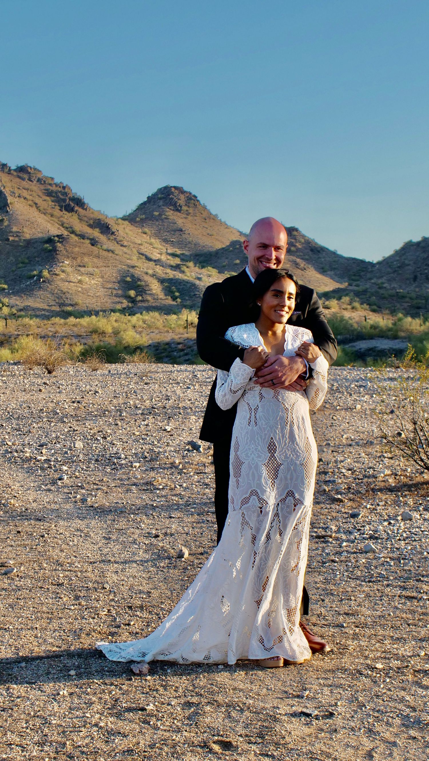 A romantic desert photoshoot featuring a couple in formal attire embracing against a backdrop of arid mountains at sunset.