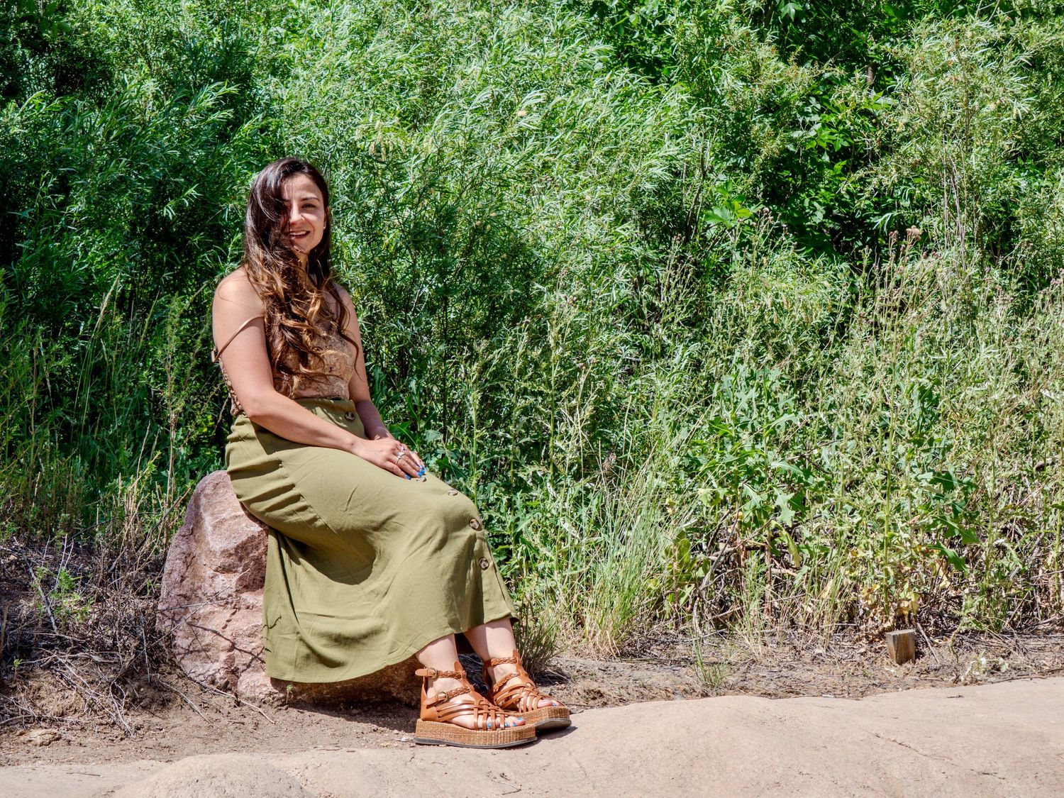 A person in a flowing olive dress sits on a rock along a nature trail surrounded by lush green foliage.