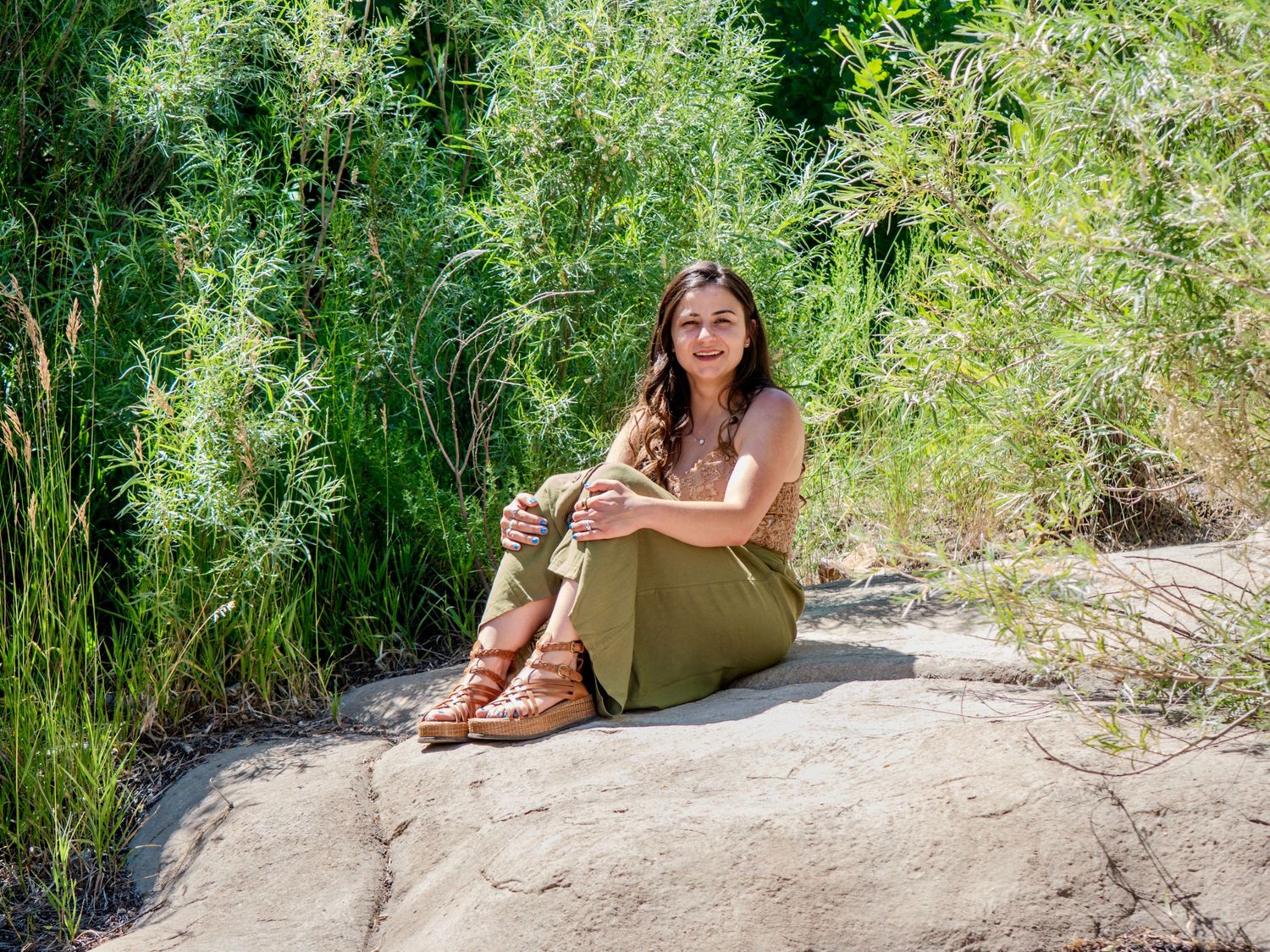 Person sitting outdoors on sandy surface surrounded by green foliage on sunny day.