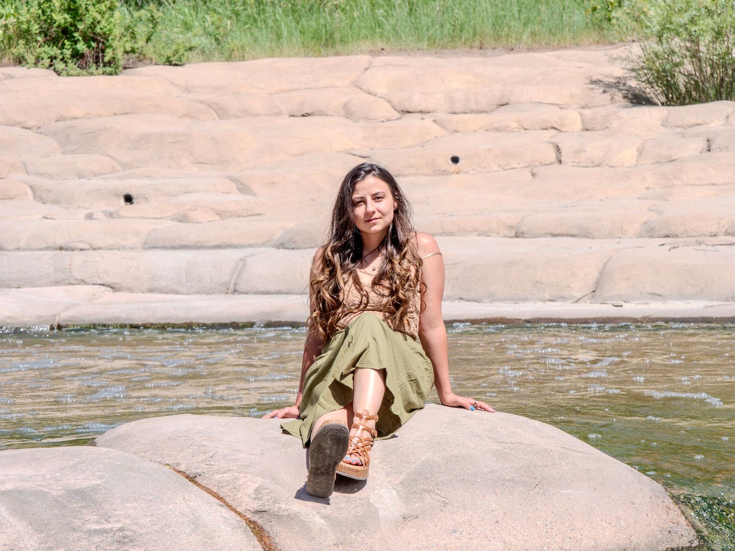Someone in a green dress sits on a large pink rock formation near a pond on a sunny day.