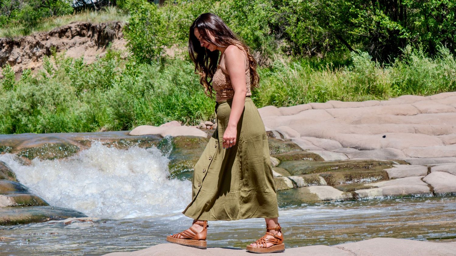 A person in olive green pants and sandals walks along rocky terrain near flowing water on a sunny day.