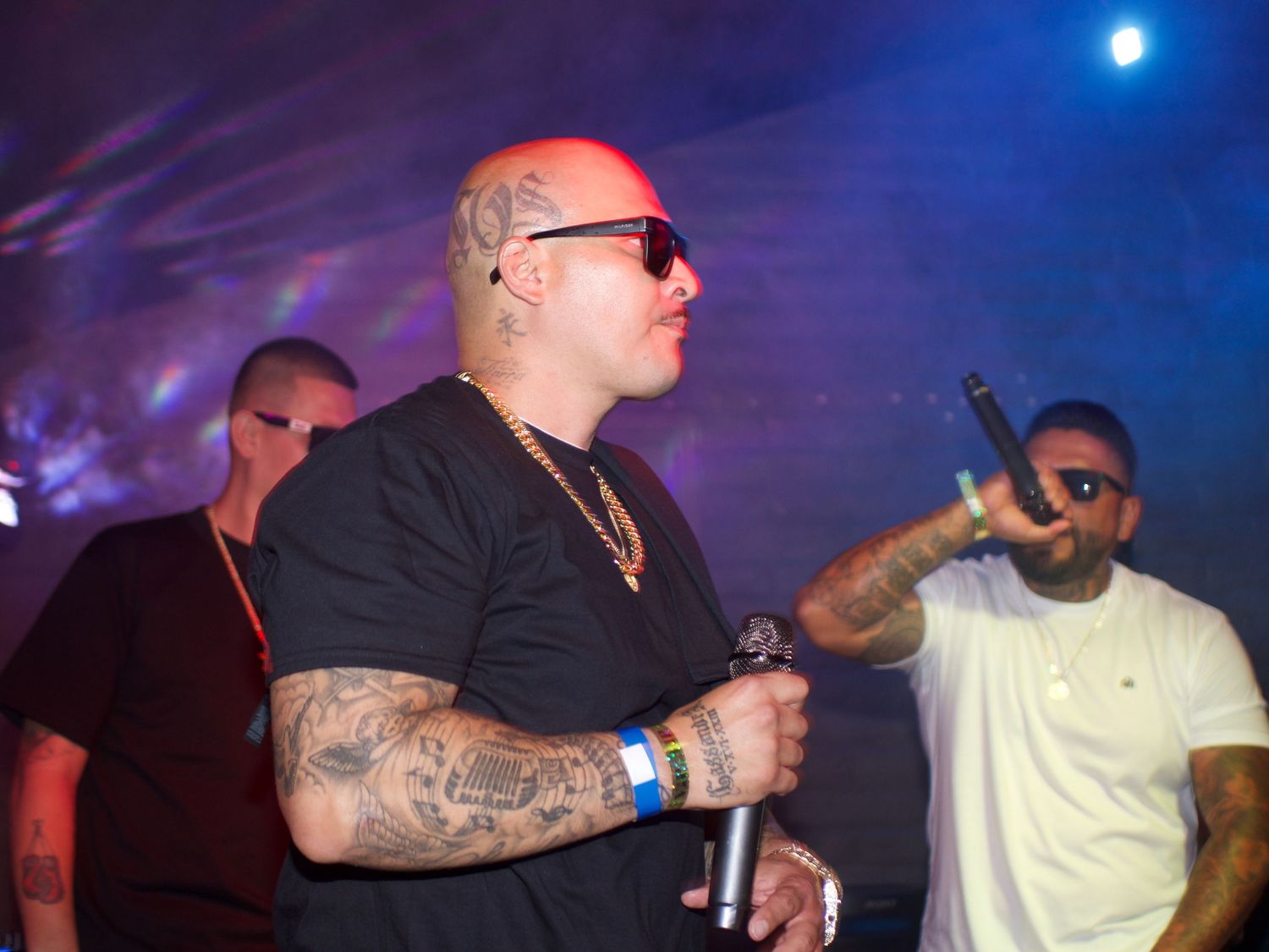 A performer with tattoos and sunglasses holds a microphone on stage with blue lighting and other performers in the background.