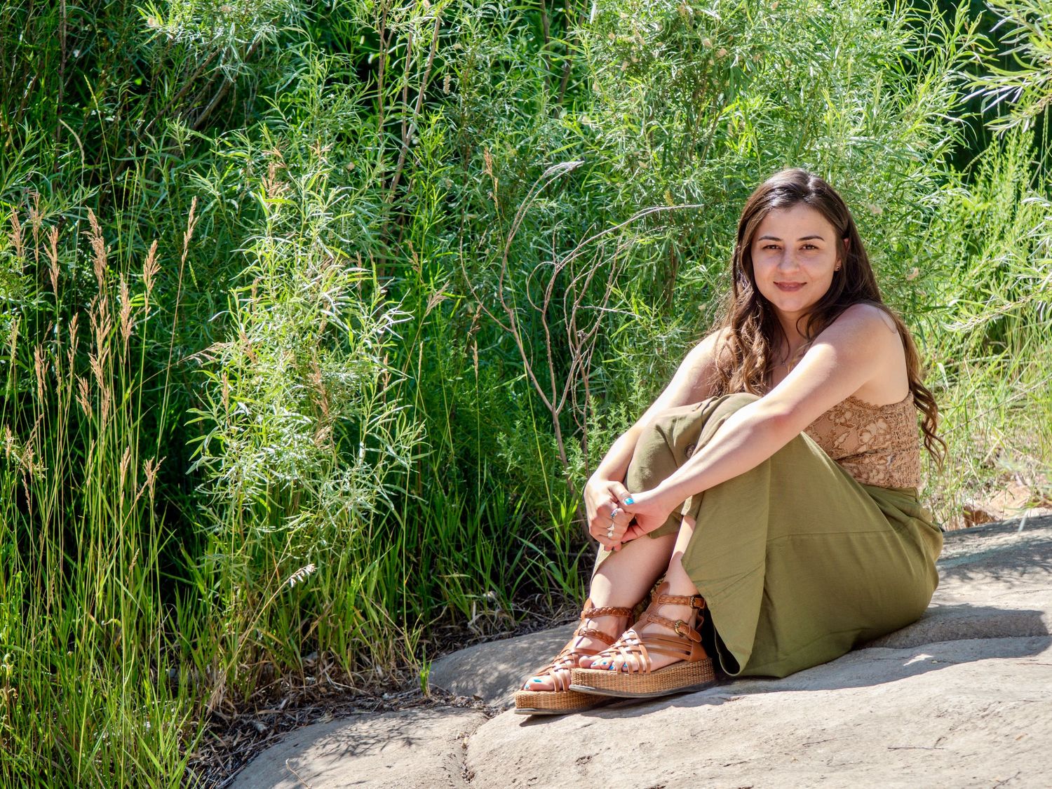 Someone in an olive green dress lounges on a large rock surface beside tall grass in a natural outdoor setting.