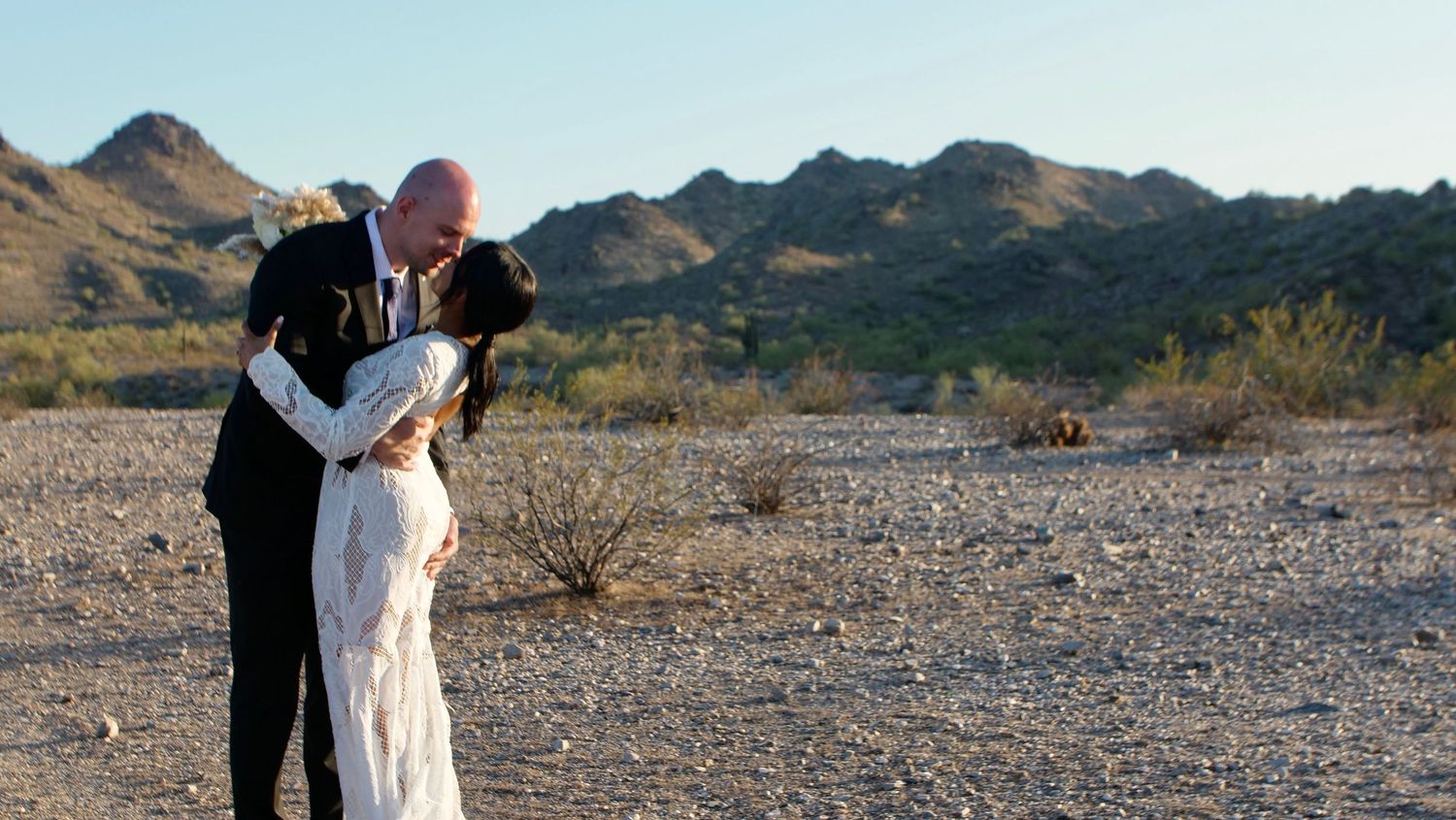 Wedding couple shares sweet moments in a desert landscape with mountains in the background during sunset.