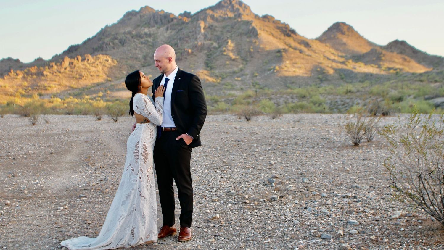 Couple in formal wedding attire poses against desert mountain backdrop at sunset.
