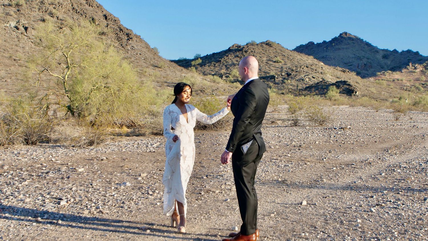A couple stands together in an outdoor desert setting with mountains and brush in the background.