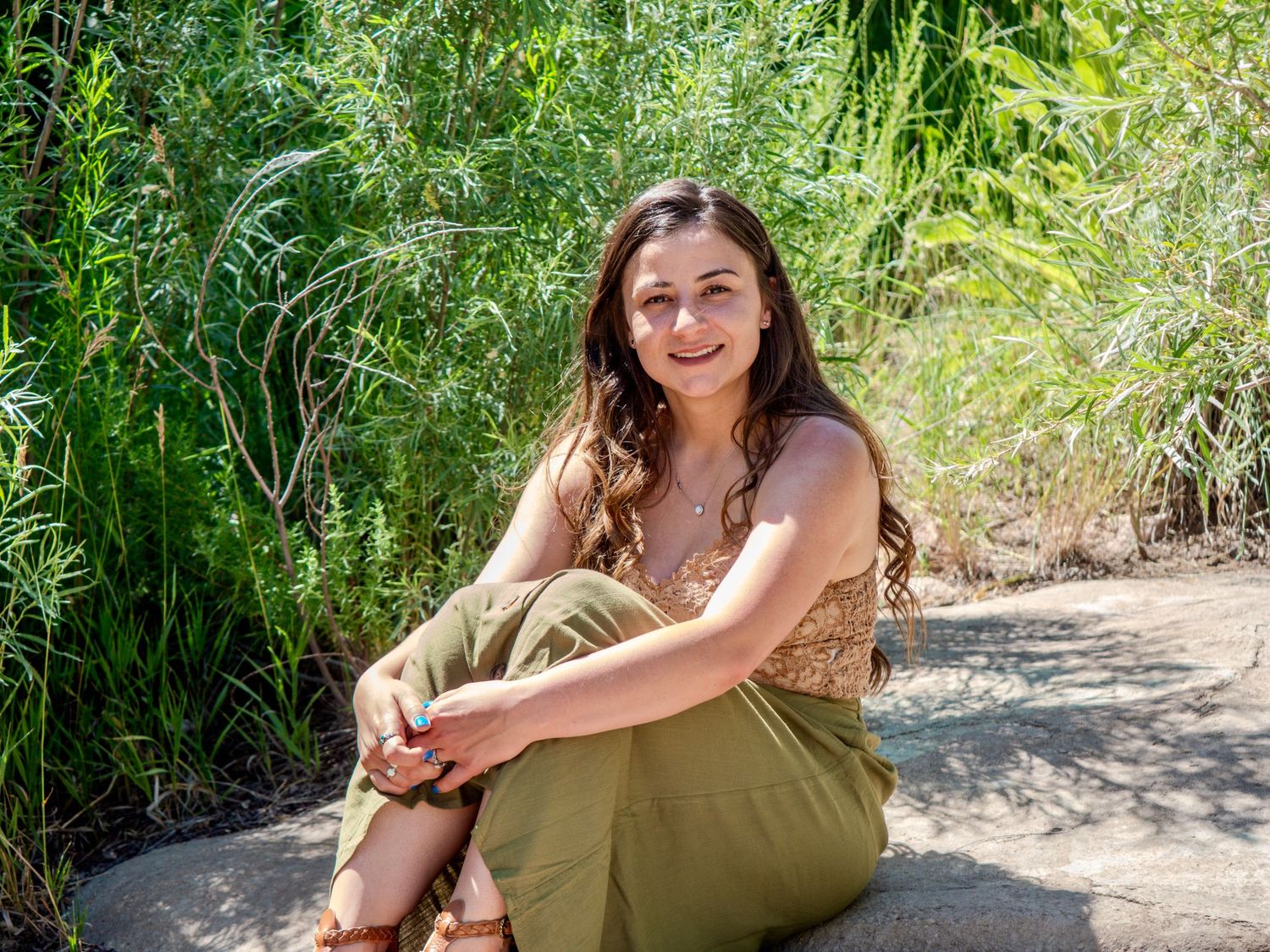 Someone in an olive green skirt and brown top sits smiling on a path surrounded by lush green plants and foliage.