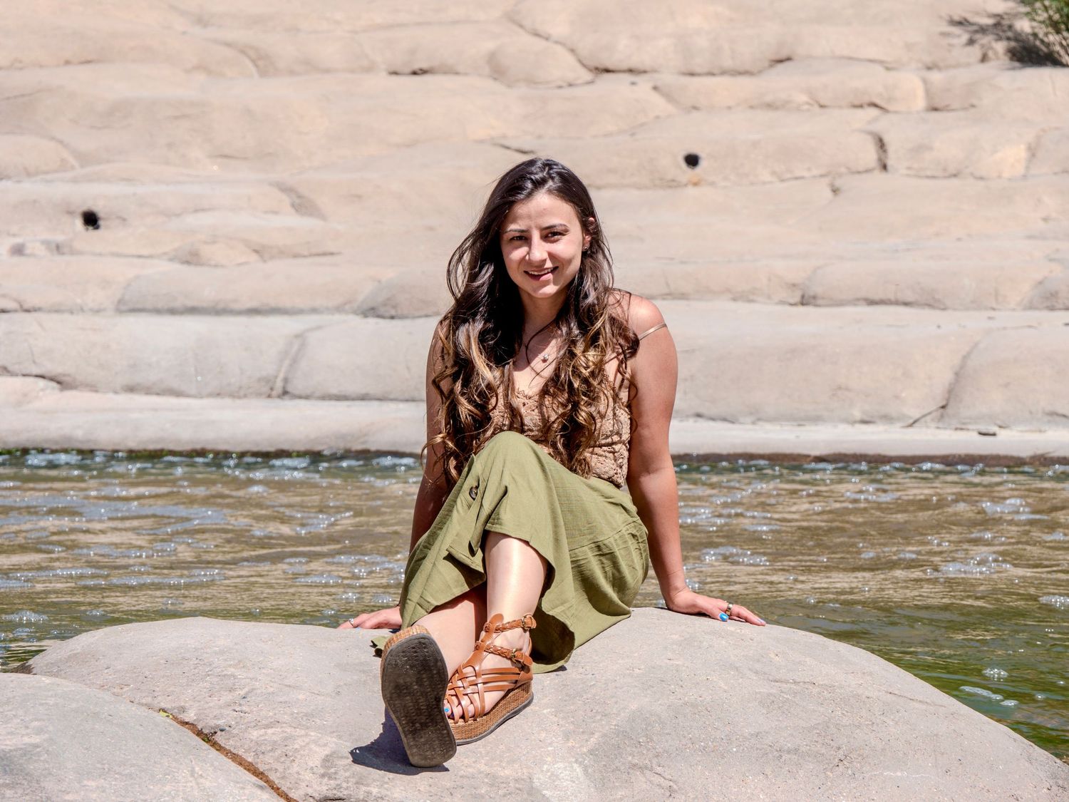 A person wearing a green dress sits on a rocky formation near a body of water on a sunny day.