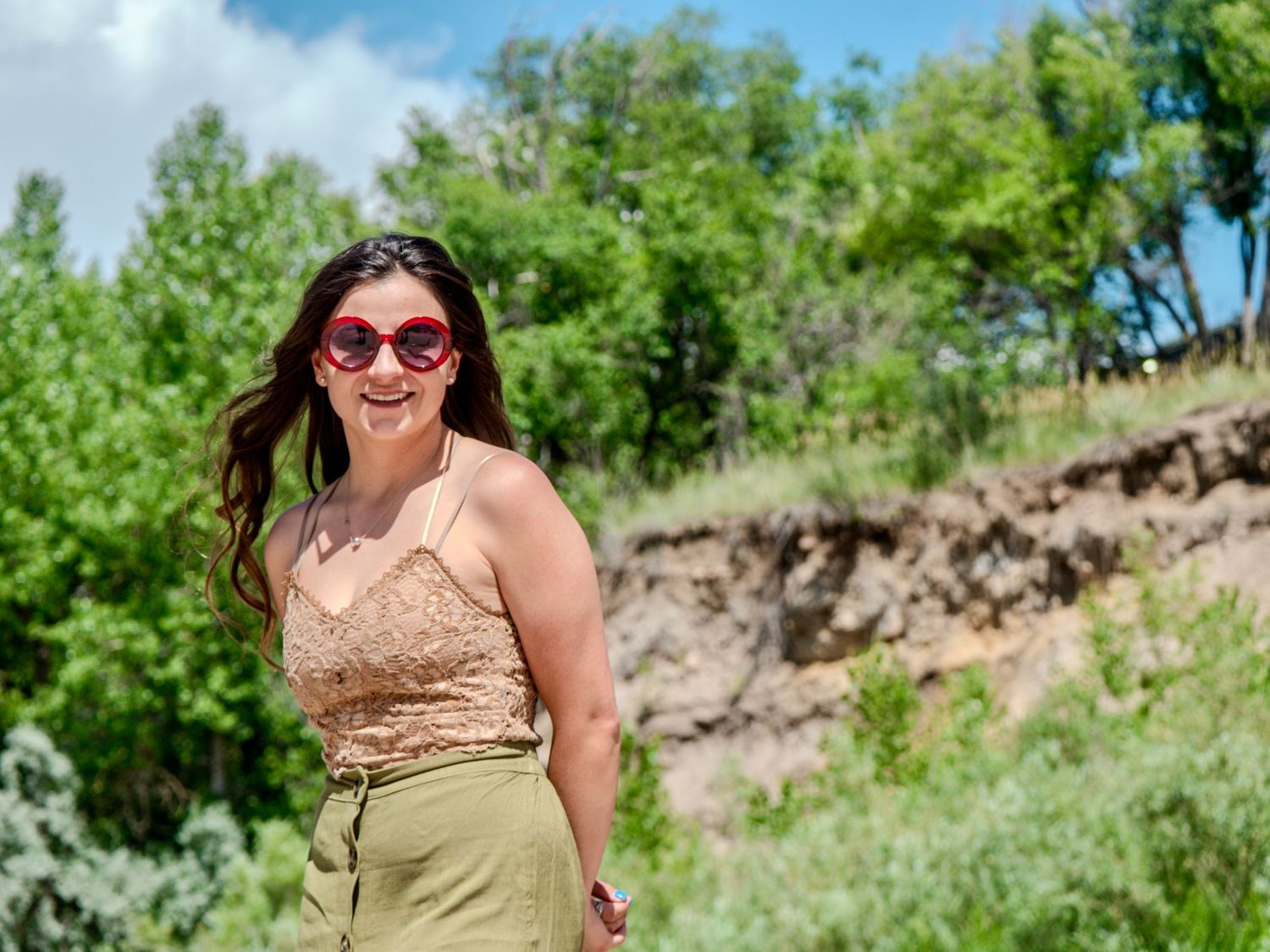 Person in sunglasses and beige top smiling outdoors with green trees and hillside in background.