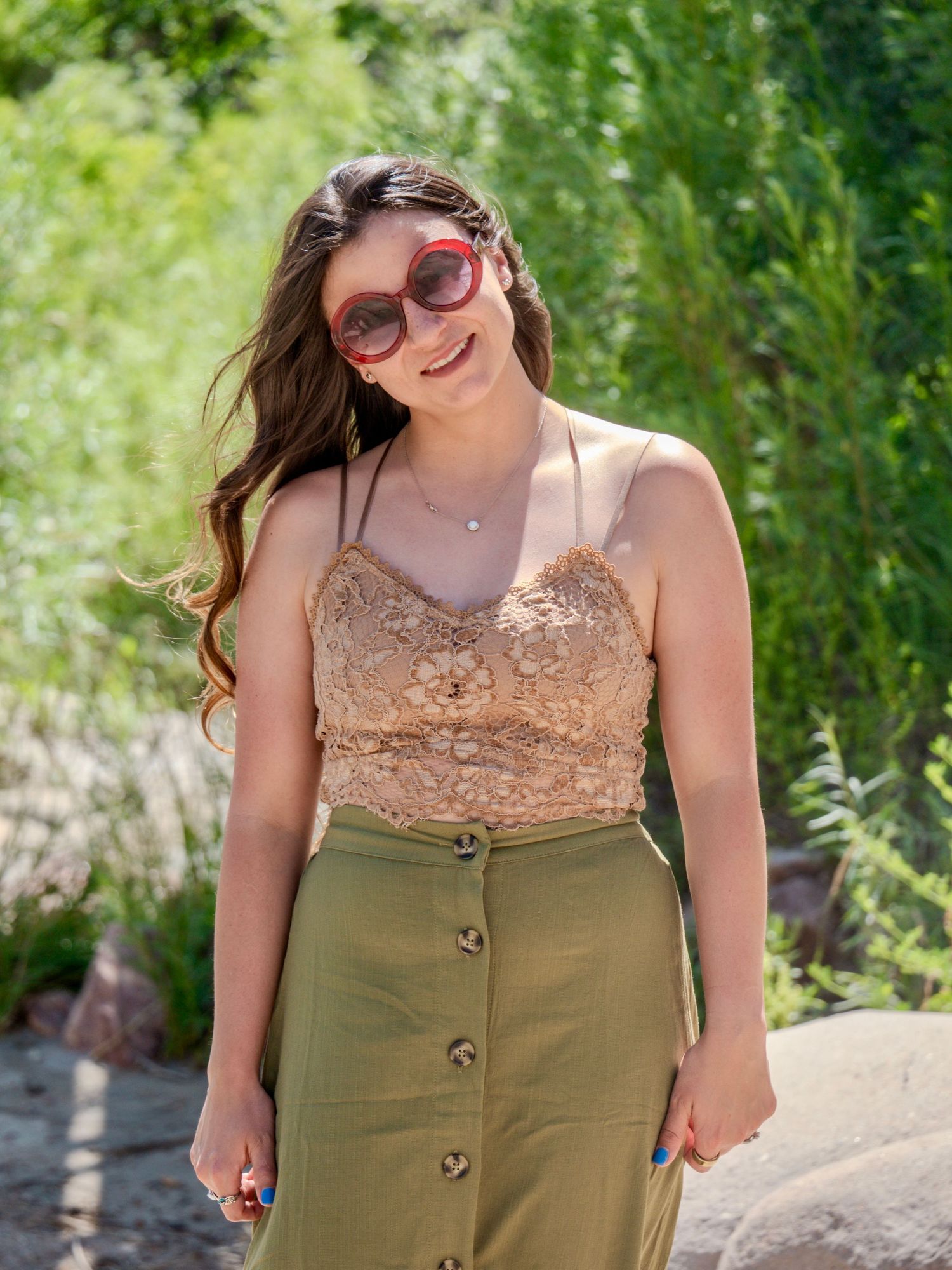 A person wearing a lacey beige top and olive green skirt poses outdoors against a natural green backdrop.