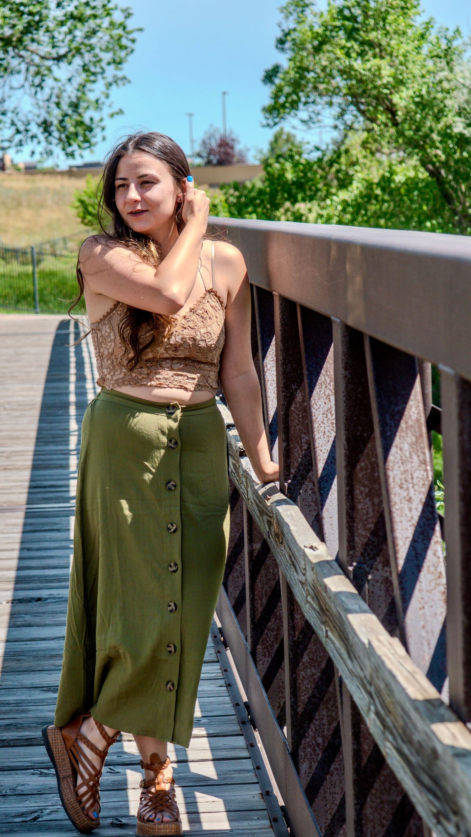 Person poses in brown top and olive buttoned skirt on wooden bridge walkway during sunny summer day.