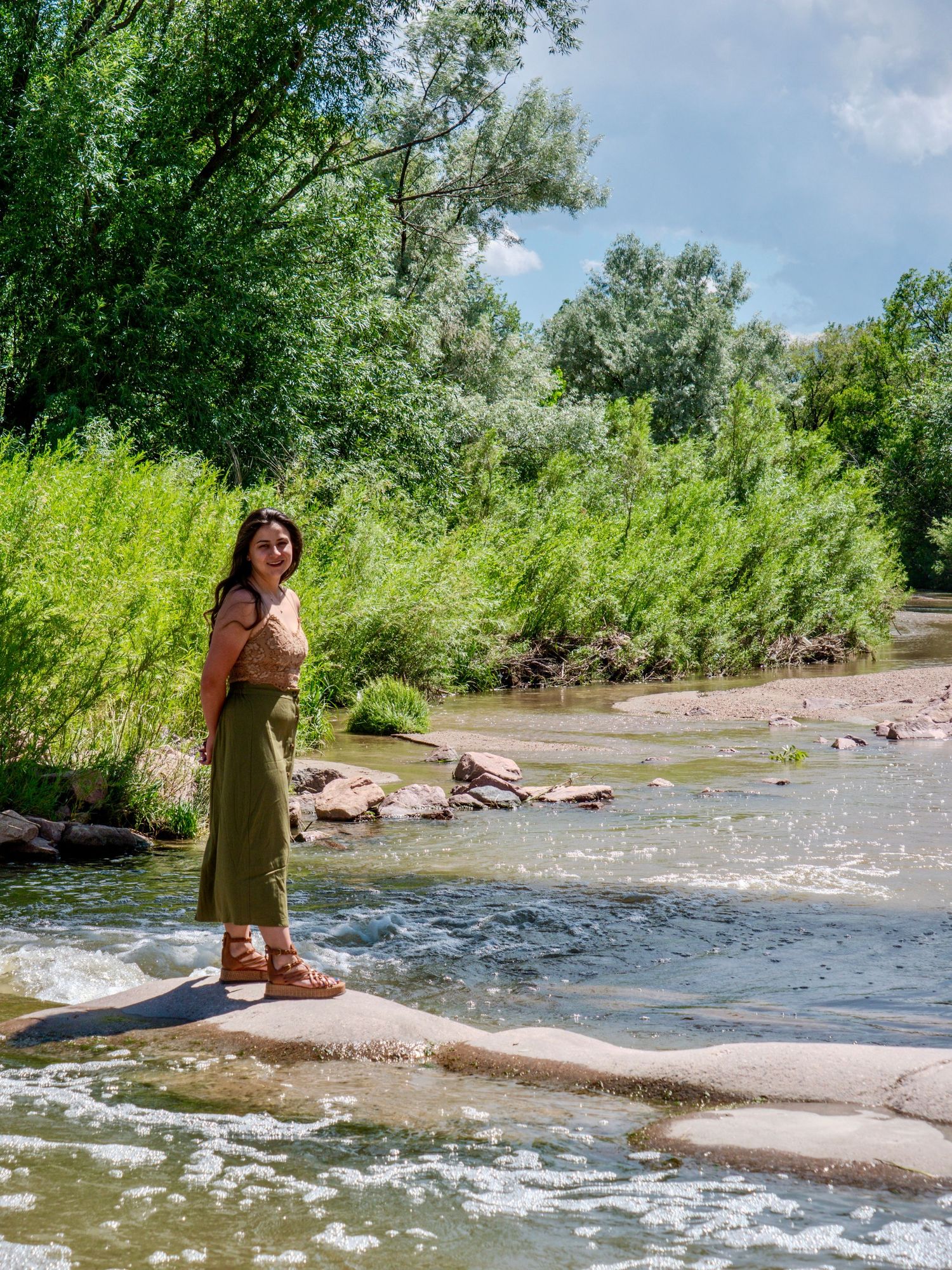 Person in a long skirt stands on rocks by a flowing river surrounded by lush greenery on a sunny summer day.