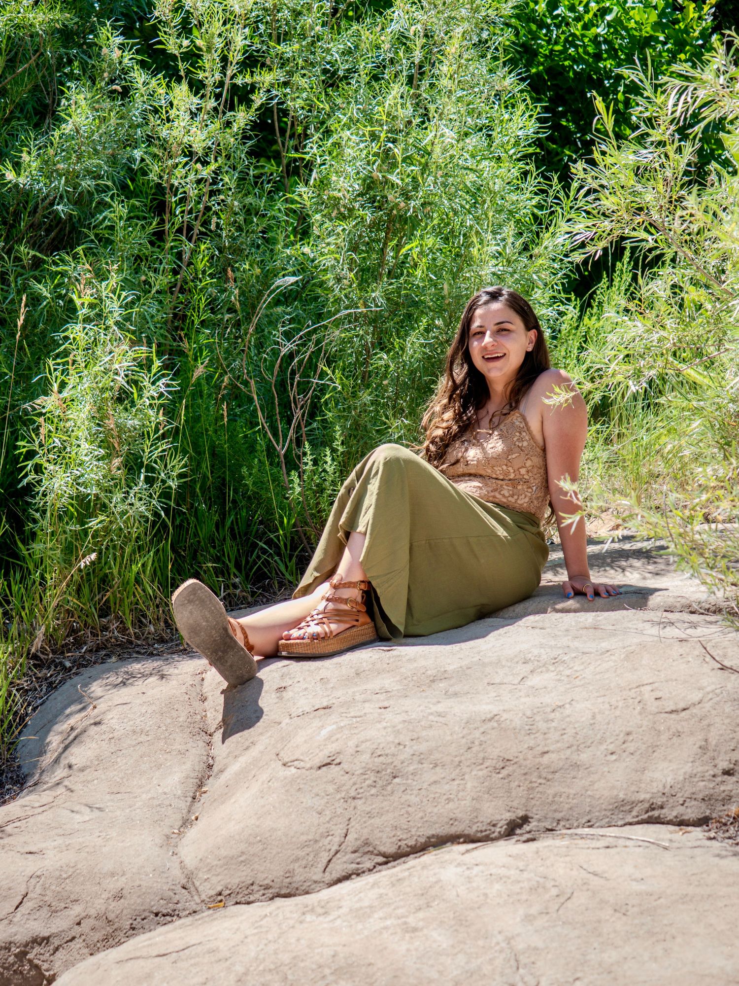 A woman sits on a large rock surrounded by lush green vegetation while wearing olive pants and a brown top on a sunny day.