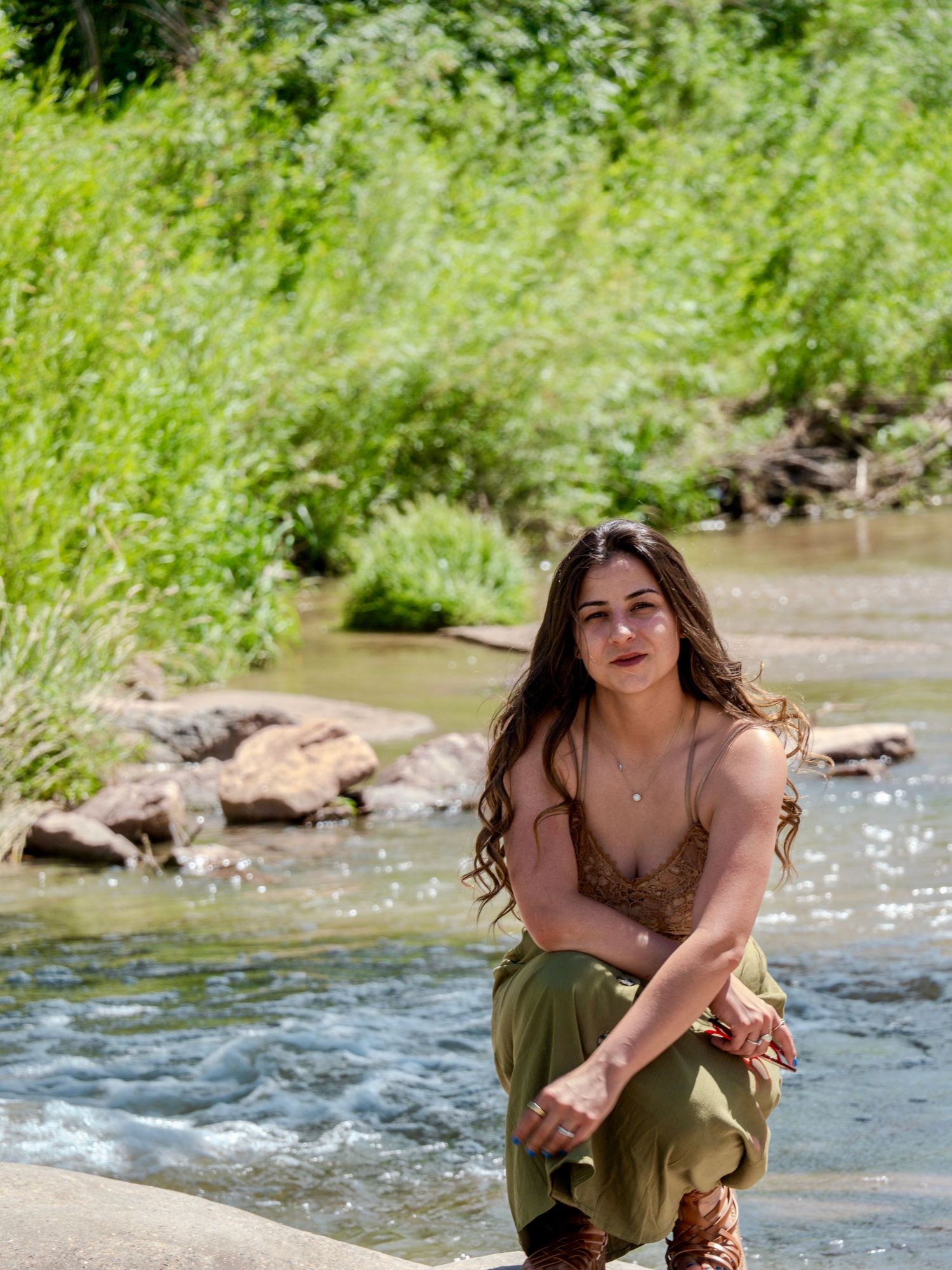 Young woman wearing an olive green dress poses by a flowing stream on a sunny summer day.