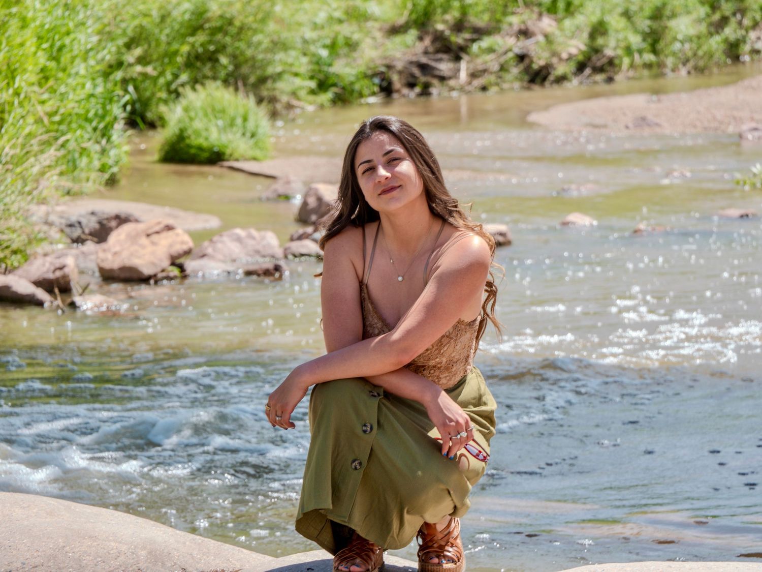 Woman in a green dress sits by a sparkling river on a sunny summer day.