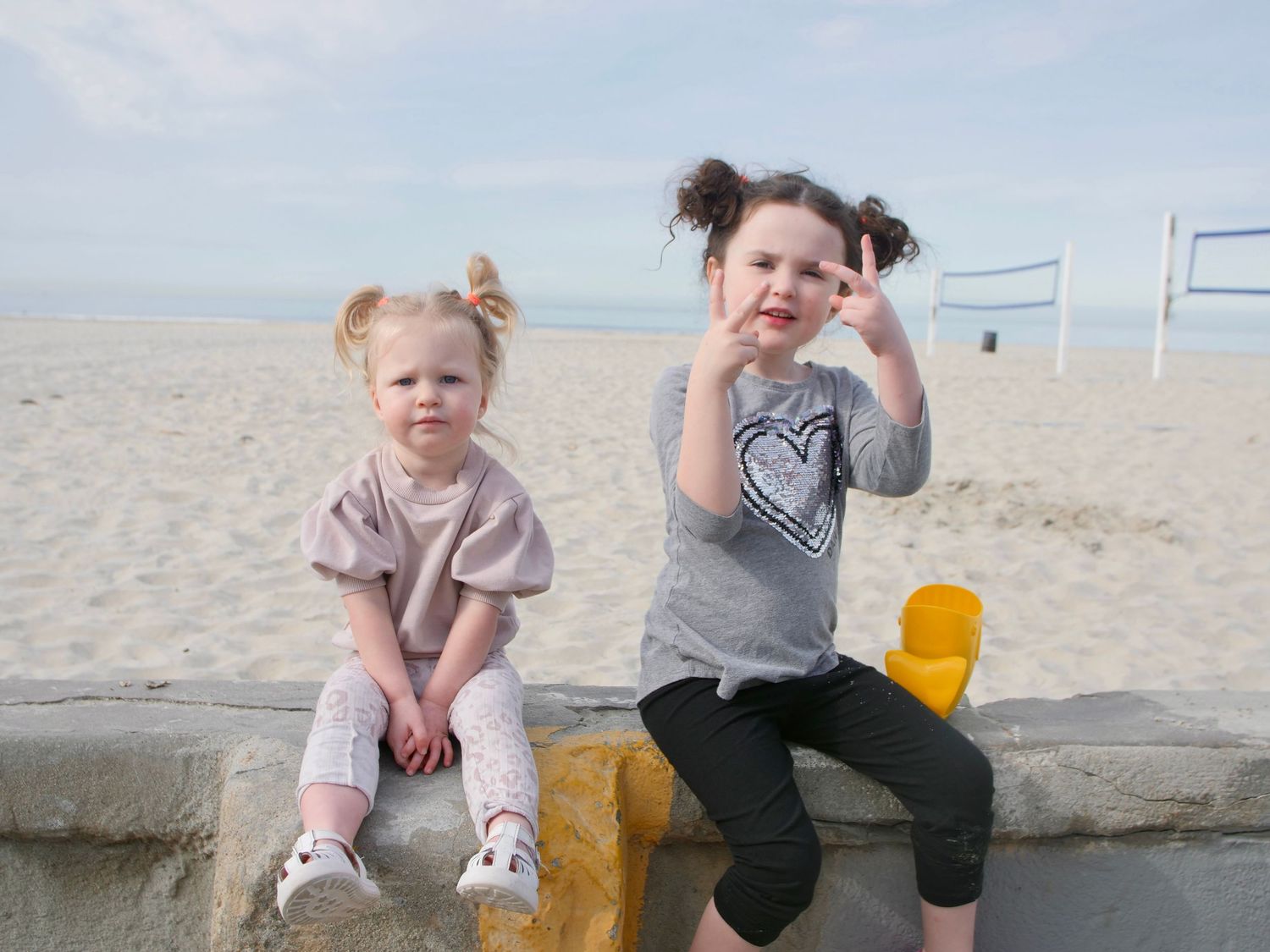 Two children sit on a concrete ledge at a sandy beach with volleyball nets visible in the background.