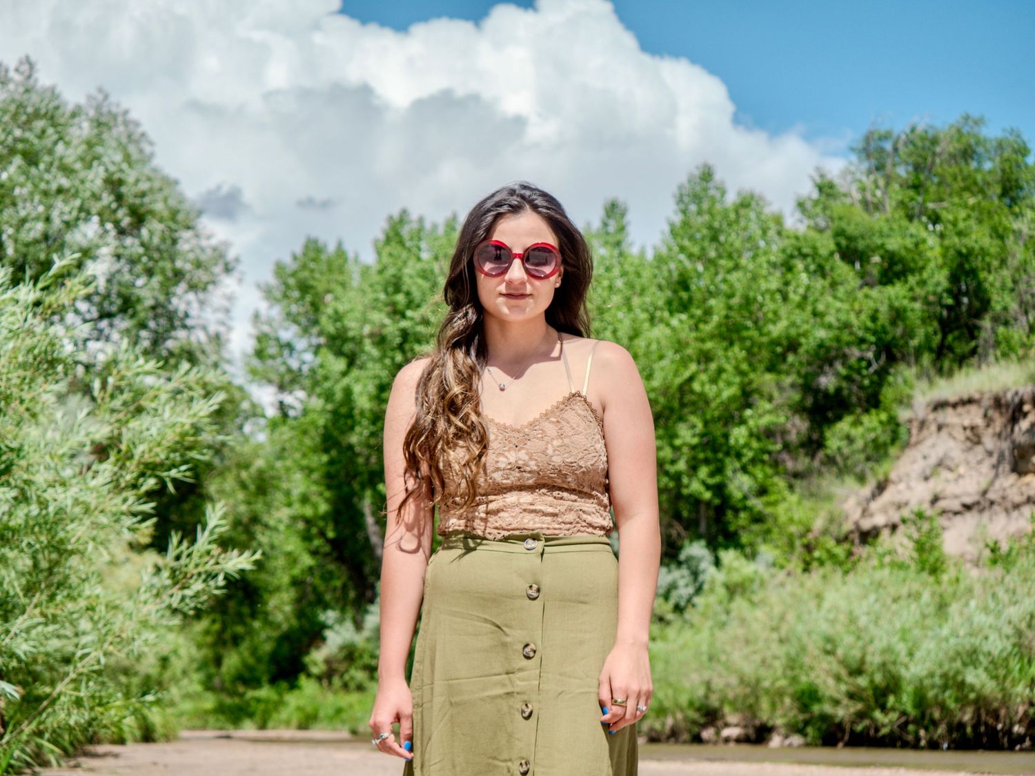 Woman in beige tank top and sunglasses standing in lush green summer garden setting.