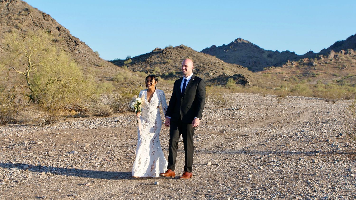 Two people pose together in formal attire against a desert mountain landscape with brown peaks and rugged terrain.
