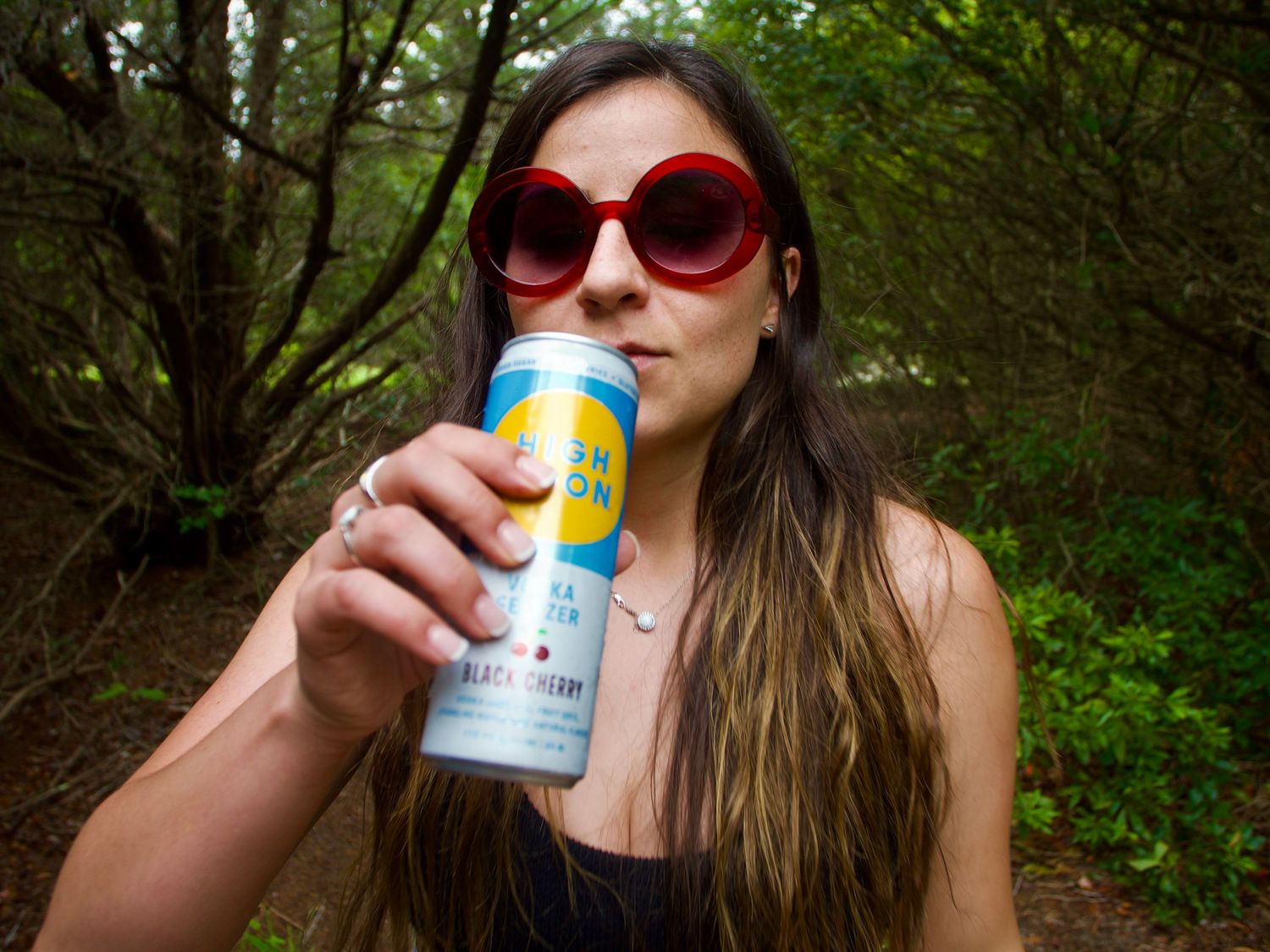 A person in sunglasses enjoying a canned beverage while sitting in a wooded outdoor setting on a sunny day.