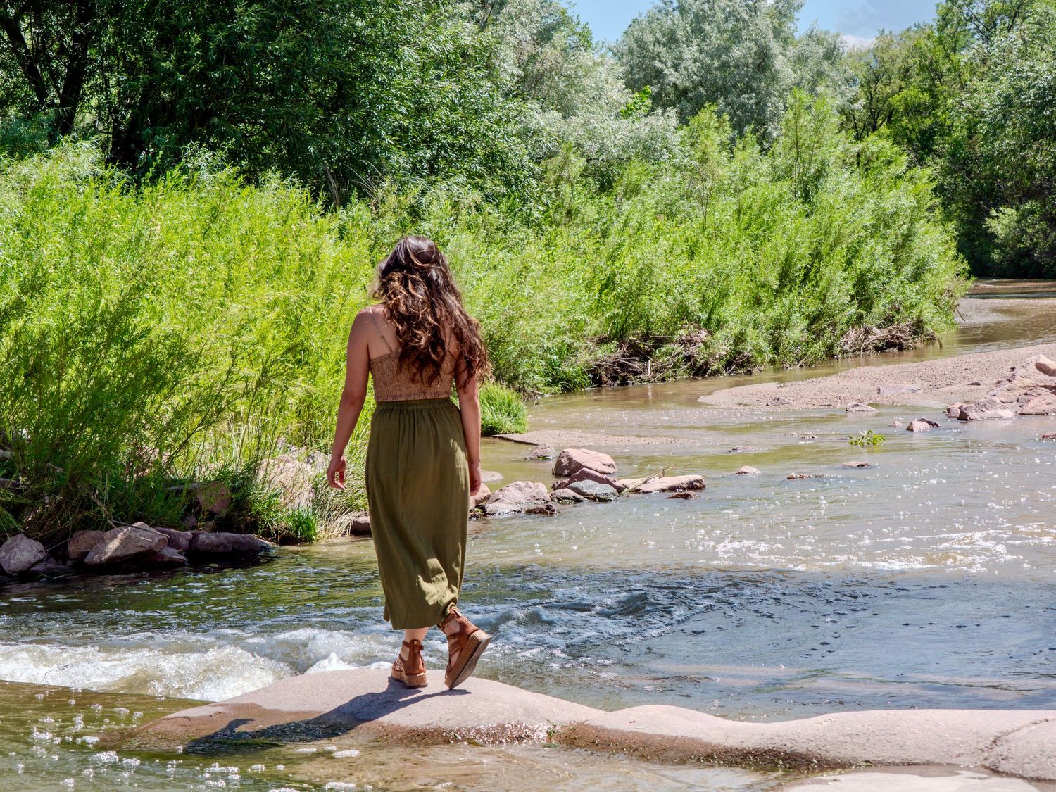 A person in a long skirt and tank top explores a shallow river on a sunny summer day, walking across red rocks.