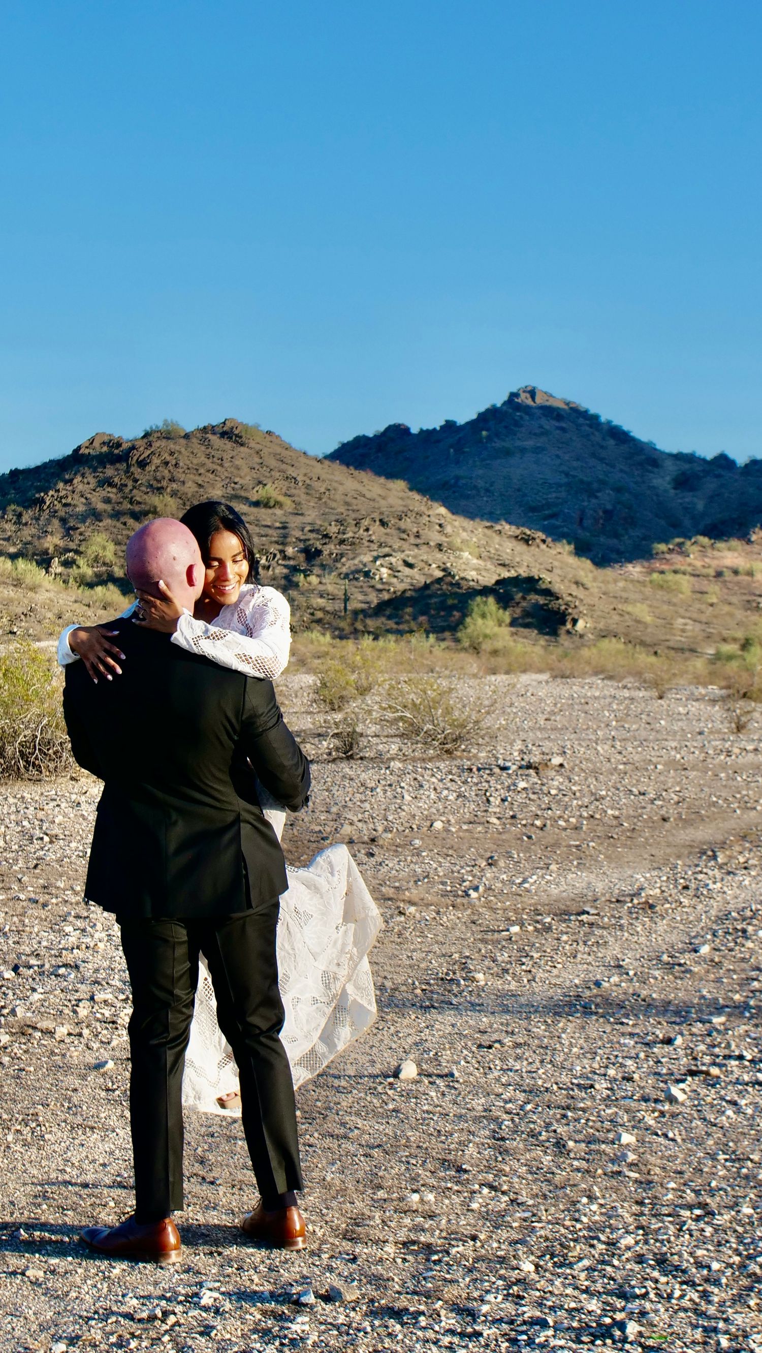 Two people in formal attire embrace on rocky desert terrain with mountains in the background.