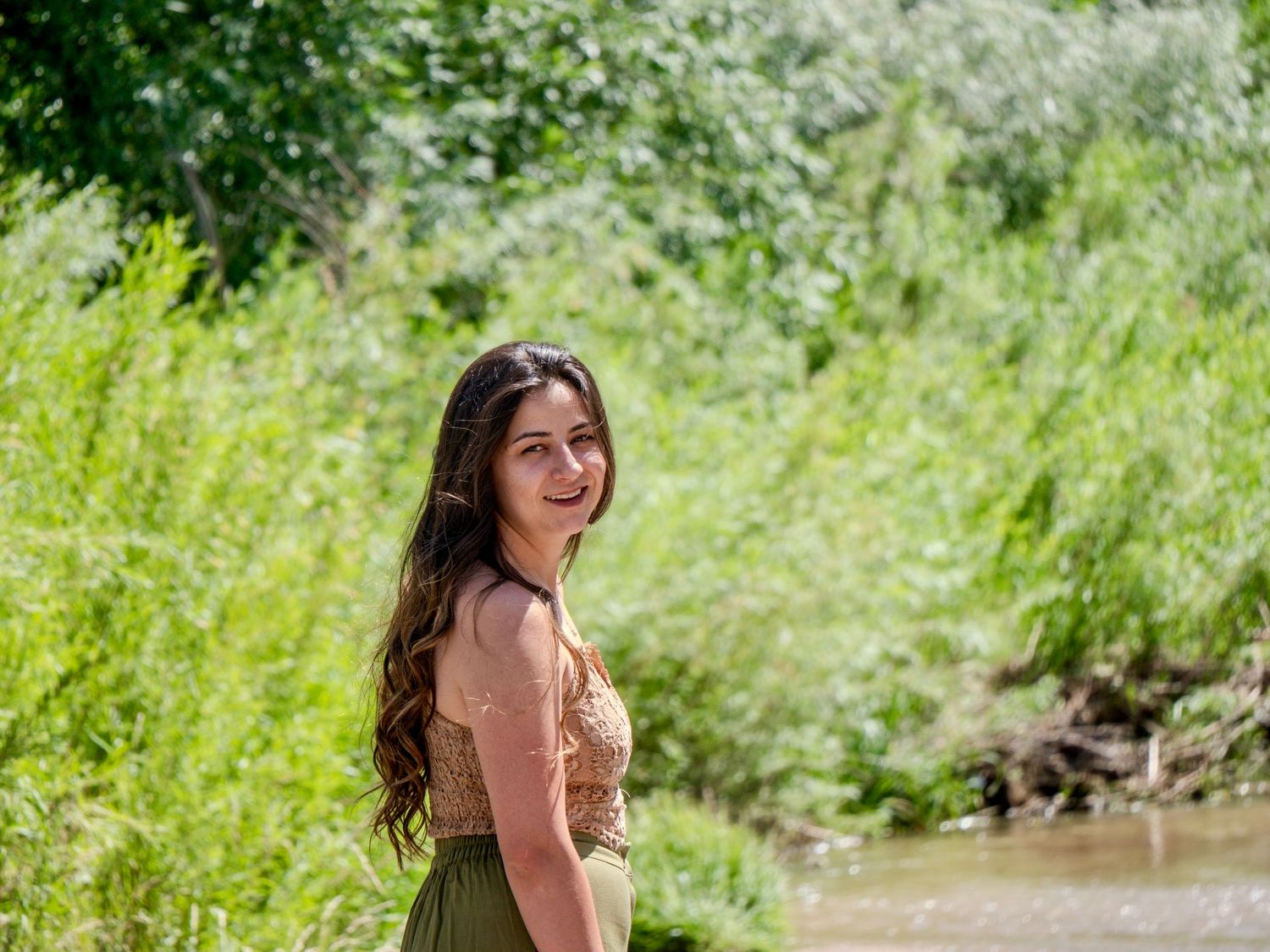 A woman in a brown dress smiles near a river bank on a sunny summer day surrounded by lush green vegetation.
