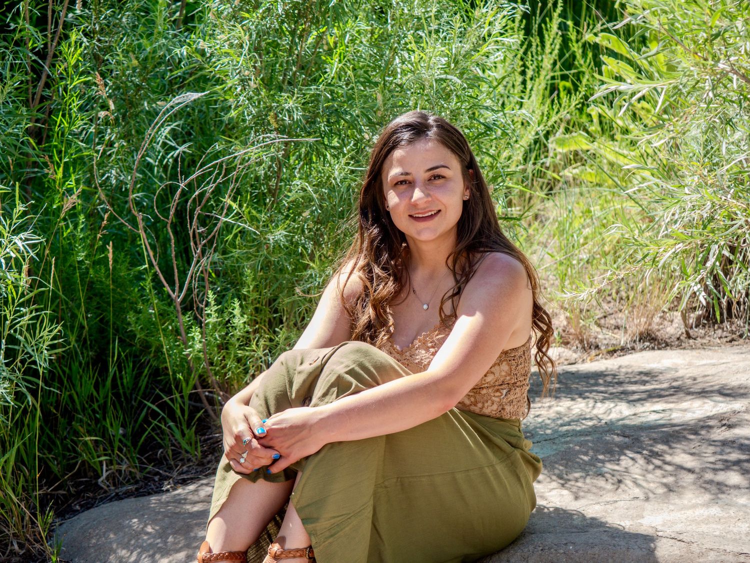 A person in an olive green dress sits casually on a dirt path surrounded by lush bamboo and greenery on a sunny day.