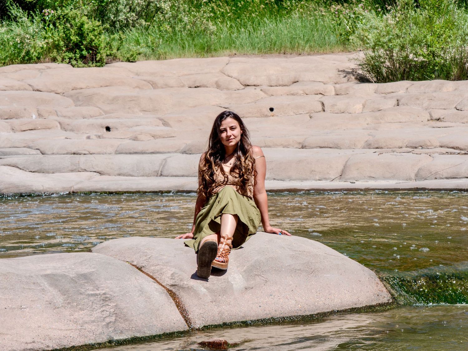 Someone sits on a large rock formation by a calm pool of water on a sunny day in nature.