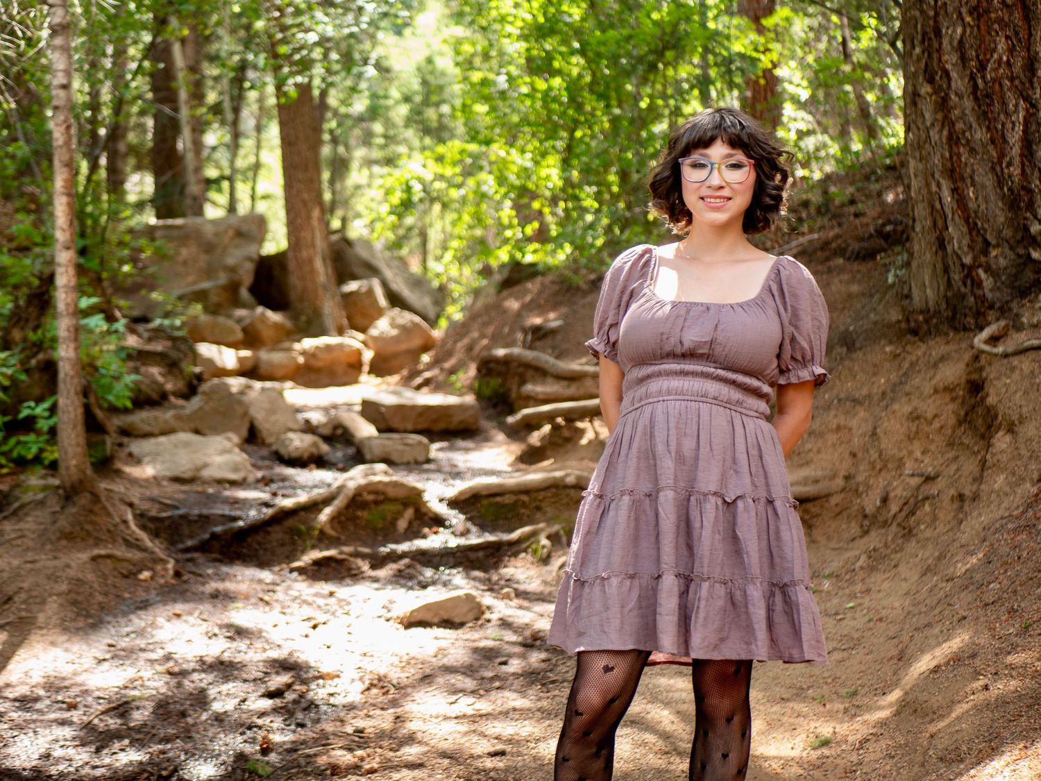 Dusty purple tiered dress worn in a sunlit forest setting with rocks and trees in the background.