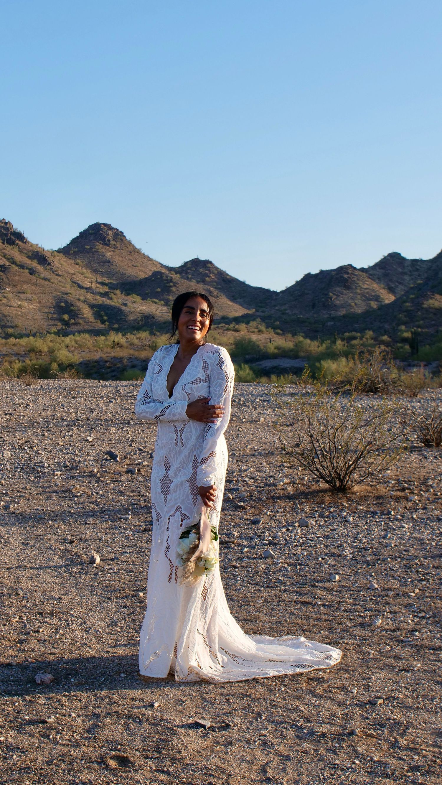 A white lace dress with train stands out against rugged desert mountains and clear blue sky in golden sunlight.