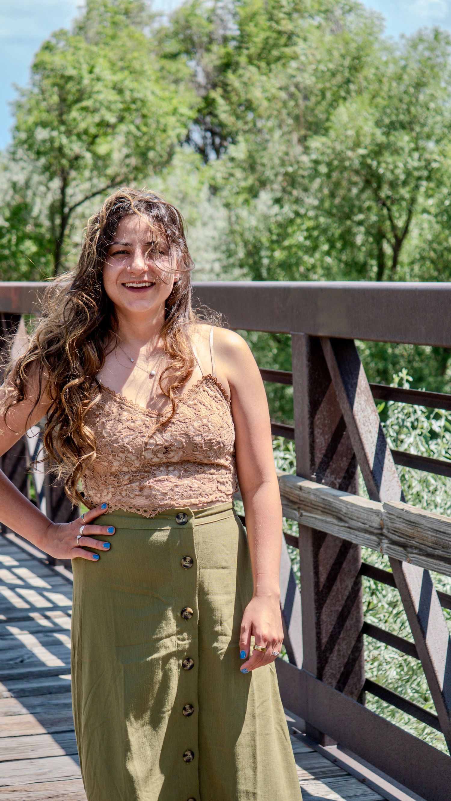 A person in a brown top and olive skirt poses on a wooden bridge in a sunny outdoor setting.