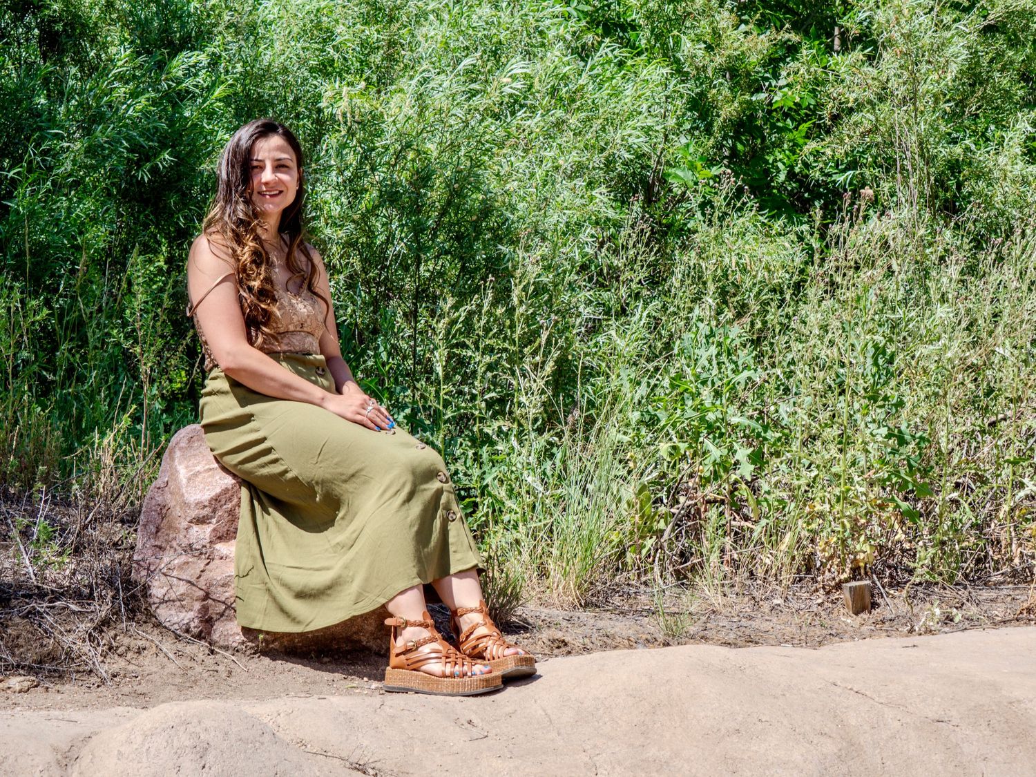 A woman in a flowy olive dress and brown sandals sits on a stone wall surrounded by lush green foliage.