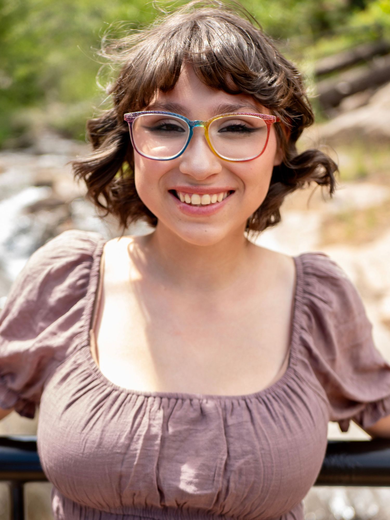 A person wearing glasses and a mauve colored top smiles brightly in an outdoor setting.