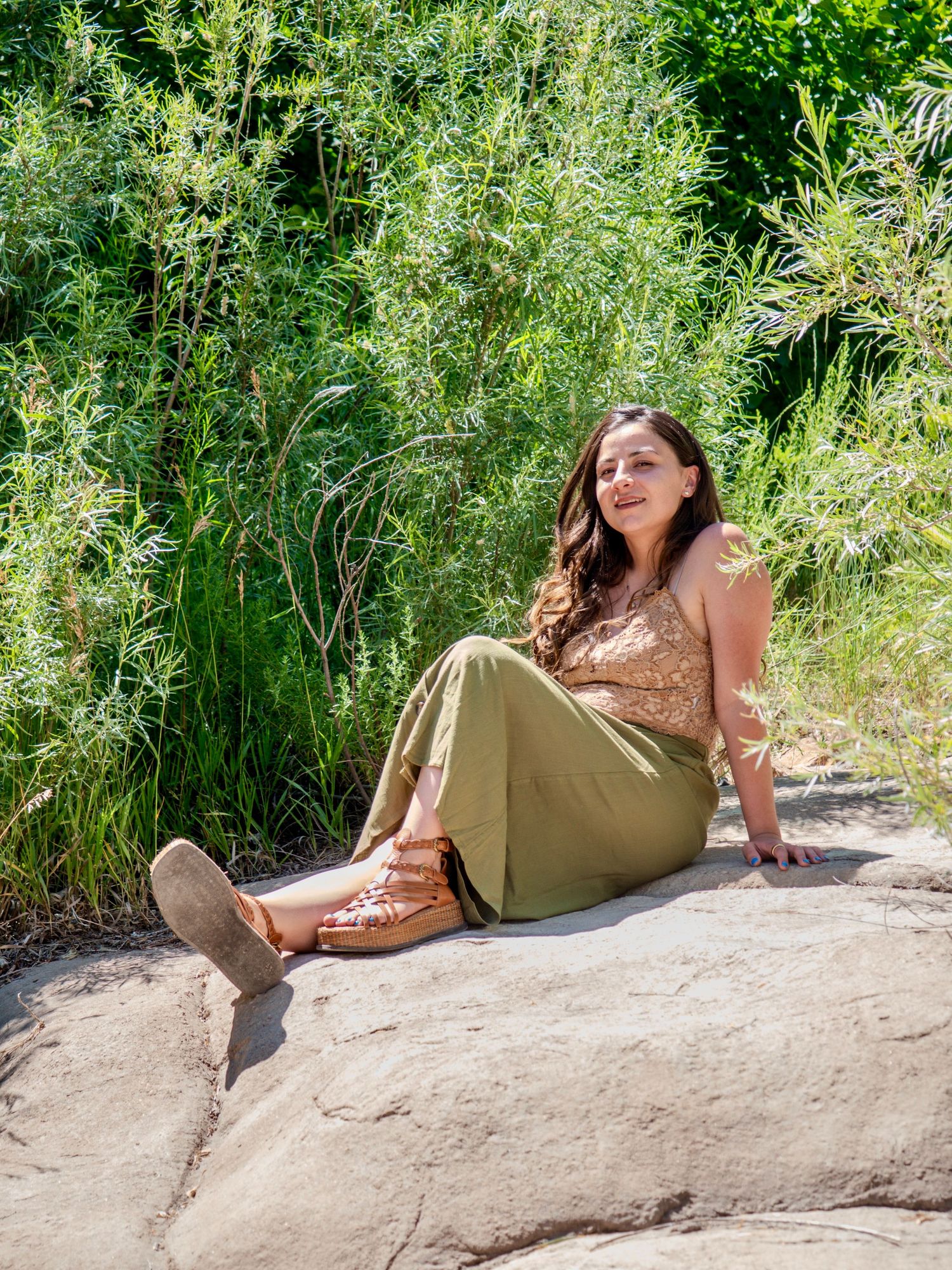 Person in brown top and olive pants relaxes on a rocky perch surrounded by tall green grasses in nature.