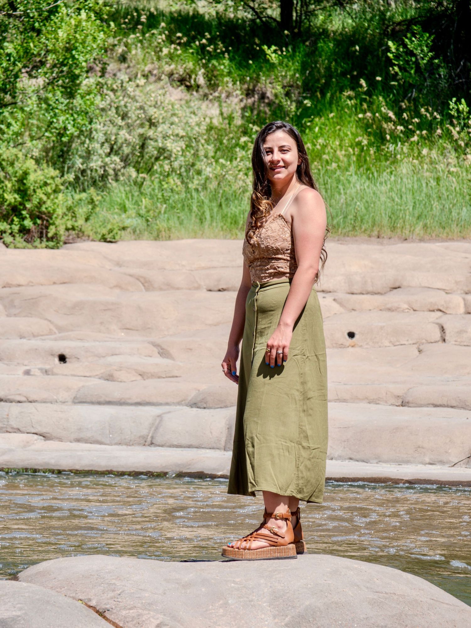 Someone poses on a rock along a riverbank wearing a long olive green dress and sandals on a sunny summer day.